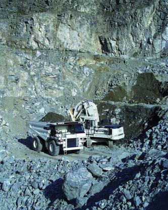 Das Bild zeigt einen Steinbruch mit mehreren Abbausohlen, in welchem graues Gestein ansteht. In der Bildmitte, auf der untersten Sohle, befinden sich zwei Fahrzeuge zur Gewinnung und Steinverladung.