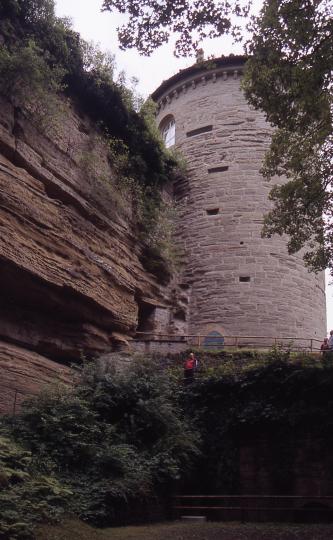 Blick auf eine hohe, rötlich braune Gesteinswand (links im Bild) sowie einen daran angebauten runden Turm aus rötlich grauem Mauerwerk (rechts). Kuppe und Fuß der Steinwand sowie das Fundament des Turmes sind von Sträuchern bewachsen.