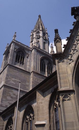 Aussenansicht einer Kirche mit Teil des Seitenschiffs, querstehendem Turmaufbau und Turm. Das Mauerwerk besteht aus rötlich grauem Gestein.