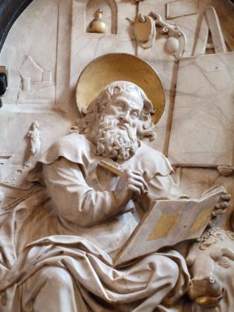 Nahaufnahme einer Heiligenfigur aus beige-grauem Stein mit gelblichen, vielleicht vergoldeten Stellen. Die Figur hält Schreibgerät und ein Buch in den Händen. Rechts unten ist ein Stierkopf zu erkennen.