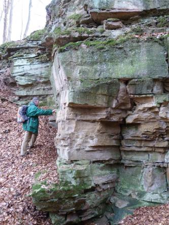 Teilansicht einer Gesteinsformation mit säulenartigem Aufbau, grünlich bis bräunlich grau. Links ist ein Hanganstieg erkennbar. Ein Mann mit Jacke und Rucksack stützt sich dort am Gestein ab.