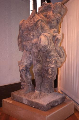 Blick auf eine ausgestellte Bildhauerarbeit aus römischer Zeit. Material rötlich graues Gestein, mit ausgebleichten und verwitterten Stellen. Die Figur soll Herkules darstellen, ihr fehlt allerdings der Kopf.