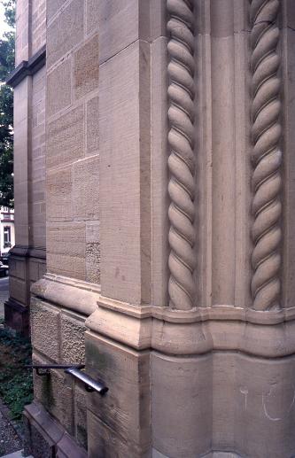 Teilansicht einer Gebäude- oder Kirchenwand aus rötlichem Mauerwerk. Rechts im Bild ist eine Säule mit Sockel und doppeltem, aus dem Stein herausgearbeiteten Zopfmuster.