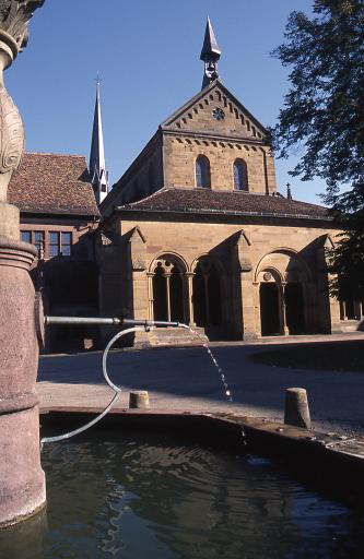 Blick auf ein Klostergebäude aus hellbraunem Mauerwerk mit Eingangshalle und spitzem Eckturm. Im Vordergrund ein Steinbrunnen.