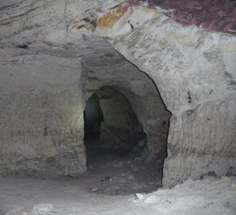 Blick in eine nur schwach erhellte Kammer aus grauem, teilweise behauenem Gestein. Ein Durchgang in der Bildmitte führt zu weiteren Kammern.