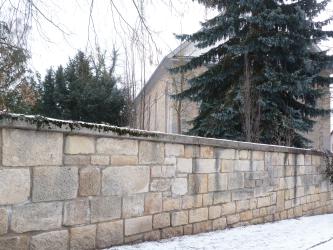 Blick auf eine hellbraune bis hellgraue Steinmauer. Im Hintergrund, hinter der Mauer und einem Nadelbaum, steht ein Kirchengebäude aus ähnlich hellem Stein. 