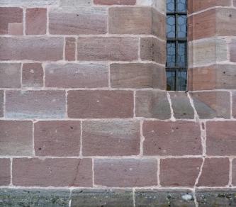 Teilansicht einer Kirchenfassade aus rötlich grauem Mauerwerk über grünlich grauem Sockel. Rechts oben ist in einer Fensterbucht ein Bleiglasfenster eingelassen.