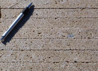 Nahaufnahme von mehreren Schichten grobkörnigem, porösem, hellbraun gefärbtem Gestein. Ein Schreibstift links oben dient als Größenvergleich.