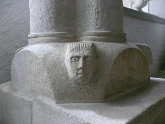 Blick auf den grauen Steinsockel einer Doppelsäule. Den Sockel schmückt eine Bildhauerarbeit (ein stilisiertes Gesicht).