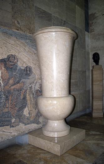 Das Bild zeigt eine sehr große, beigefarbene Vase. Die aus drei unterschiedlichen Teilen (Fuß, rundes Mittelstück und trichterförmige Öffnung) bestehende, marmorierte Vase steht auf einem niedrigen Podest.