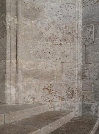 Teilansicht einer rötlich grauen Steinwand sowie flachen Treppenstufen. Die Wand knickt links und rechts ab, die Treppe verläuft davor.