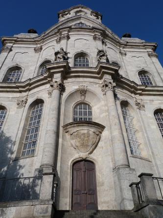 Blick auf eine mehrstöckige Gebäudefassade im barocken Stil, aus hellgrauem bis bräunlichem Stein, mit Eingangsportal, Wappen, hohen vergitterten Fenstern und durchgehenden Simsen zwischen den Stockwerken.