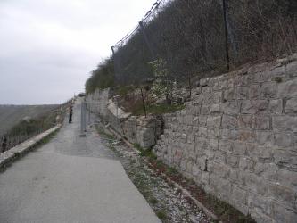 Seitlicher Blick auf eine teilweise eingestürzte Stützmauer an einem Berghang. Der Hang steigt nach rechts hin an. Links schließen sich ein Fahrweg  sowie Rebanlagen an.