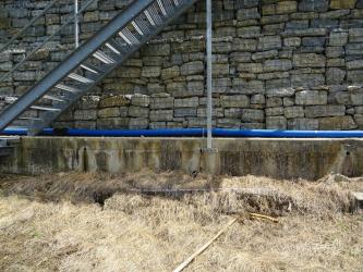 Unterhalb dem Betonsockel einer mit Gittern gesicherten Steinmauer ist eine horizontal im Boden verlaufende Spalte zu erkennen. Auf dem Sockel liegt ein blaues Leitungsrohr. Links ist ein Teil einer metallenen Aufstiegstreppe zu sehen.