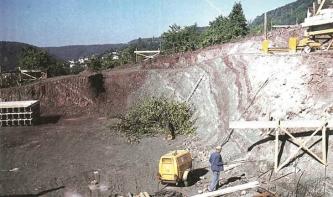 Das Bild zeigt eine Grube, in der sich ein Mensch und Geräte befinden. An den Seitenwänden steht rötliches bis graues Gestein an. Im Hintergrund bewaldete Berge.