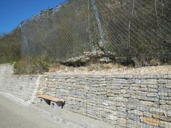 Seitlicher Blick auf einen durch eine Mauer gestützten und mit Büschen bewachsenen Hang. Nach rechts hin sind die Mauersteine durch Gitter gesichert. Darüber, zur Bergseite, ist ein Schutzzaun aufgestellt. Ein paar größere Steinblöcke liegen darin.