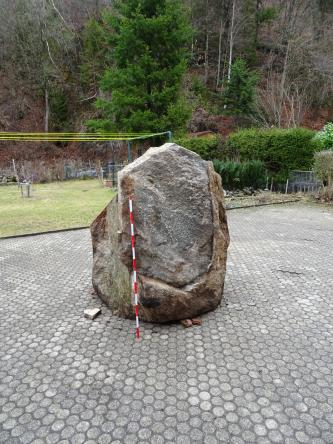 Das Foto zeigt einen einzelnen großen Felsblock. Der Block steht auf einem gepflasterten Platz. Im Hintergrund ist ein steiler Waldhang zu sehen.
