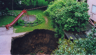 Blick von oben auf einen tiefen Krater, entstanden im Garten vor einem Haus. Der Bereich um den Erdfall ist abgesperrt. Neben dem Krater steht eine Kinderrutsche.