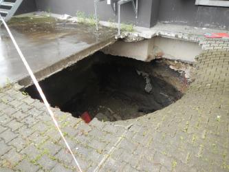 Erdfall bei einem ehemaligen Einkaufsmarkt in Schwenningen Zu sehen ist die Ecke einer Gebäudewand, davor ein Boden mit Pflastersteinen. In der Bildmitte ist der gepflasterte Untergrund eingestürzt. Ein großes Loch gibt den Blick auf das Gebäudefundament und das Erdreich unter den Pflastersteinen frei.