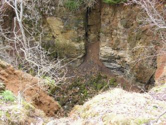 Blick von oben in den Krater einer Doline. Die hintere Wand besteht aus rötlich braunem bis grünlichem Felsgestein, das in der Mitte einen schmalen Kamin aufweist.