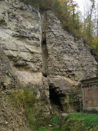 Blick auf eine hohe, nach links hin aufsteigende Felswand. Im graugelben Gestein ist ein fast durchgehender, senkrecht verlaufender Kaminschlot erkennbar. Am unteren Ende öffnet sich ein Hohlraum.